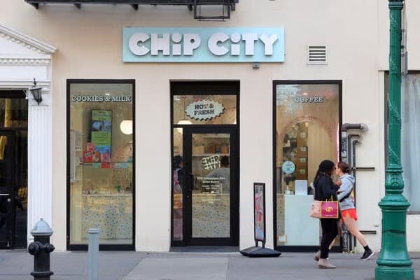 A Chip City cookie shop with a white illuminated sign above glass doors on a city sidewalk, with a pedestrian walking past.