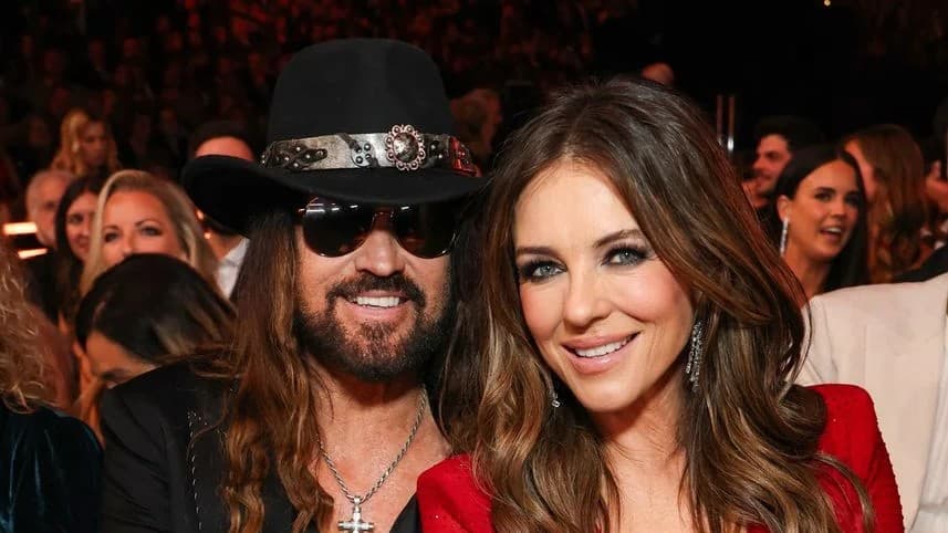 Billy Ray Cyrus, in a black hat and sunglasses, seated beside Elizabeth Hurley in a red dress at an awards show.